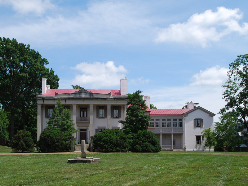 A large historic mansion with a red roof and multiple chimneys, surrounded by green lawn and trees under a blue sky.